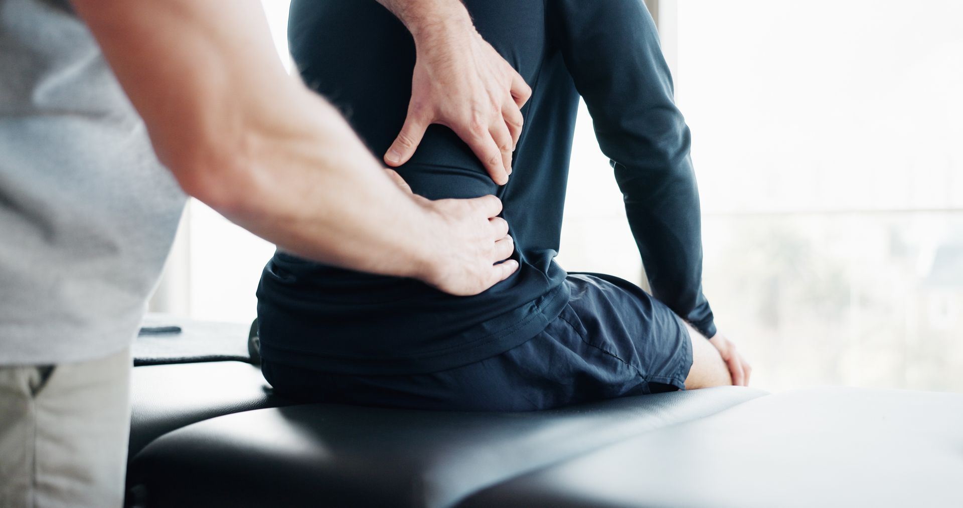 A person holding their lower back while sitting on a treatment table, with a therapist’s hands supporting them.