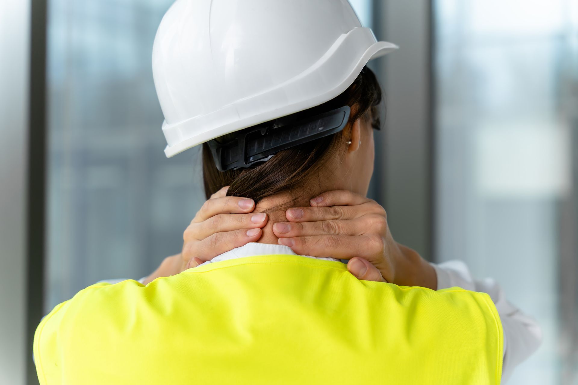 Construction worker holding neck in pain, wearing white hard hat and neon yellow safety vest