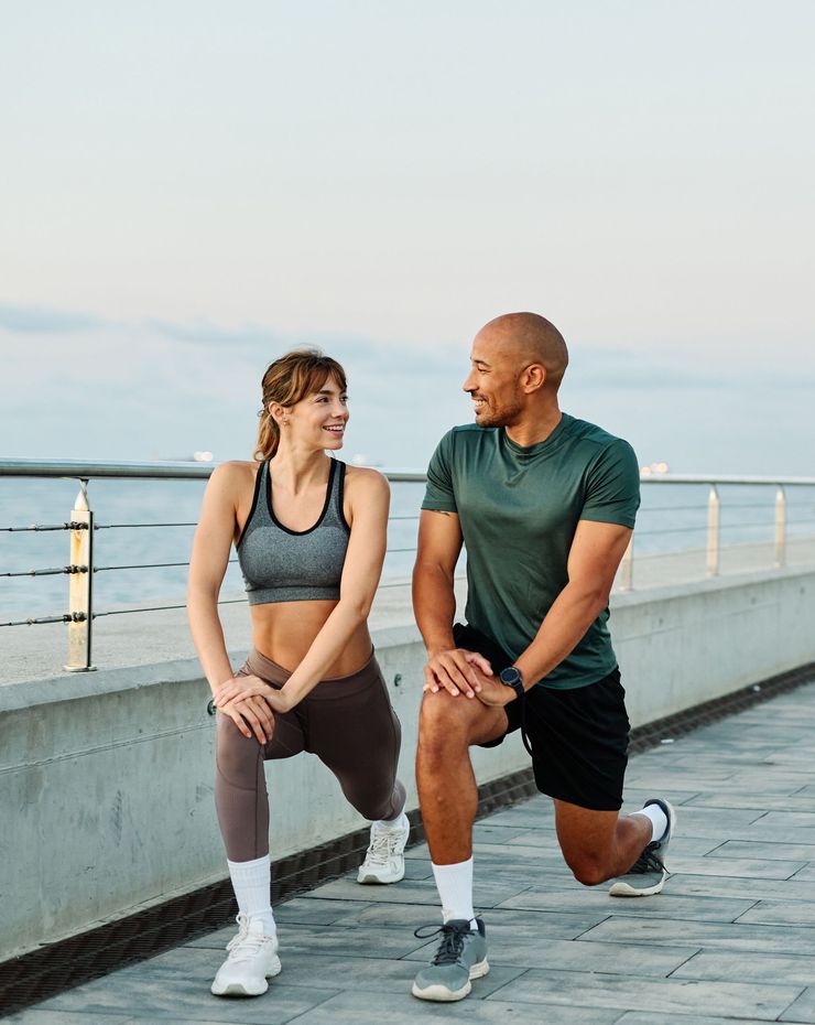 Two people lunge on a waterfront boardwalk at sunset, smiling and facing each other.