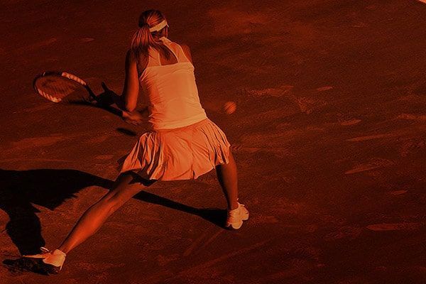 Tennis player in white outfit lunging for a shot on a red clay court under warm lighting