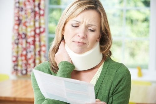 Woman wearing a neck brace reads a document at home, looking concerned.