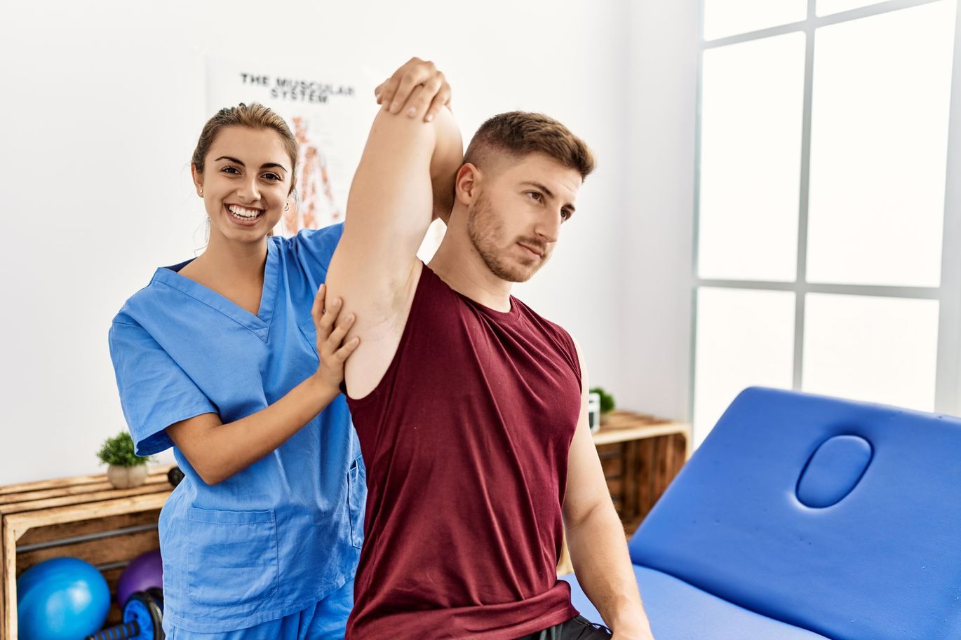 Physical therapist assisting a seated man with a seated arm stretch in a bright clinic room
