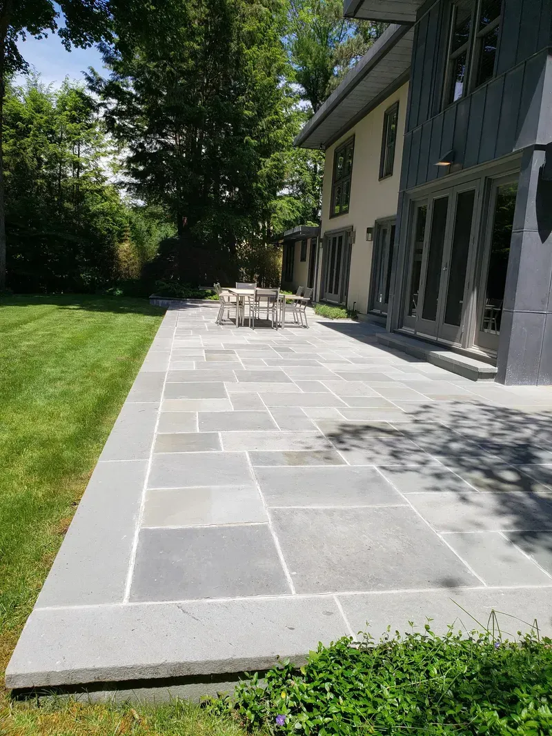 Blue stone patio next to a house with a table and chairs, surrounded by grass and trees.