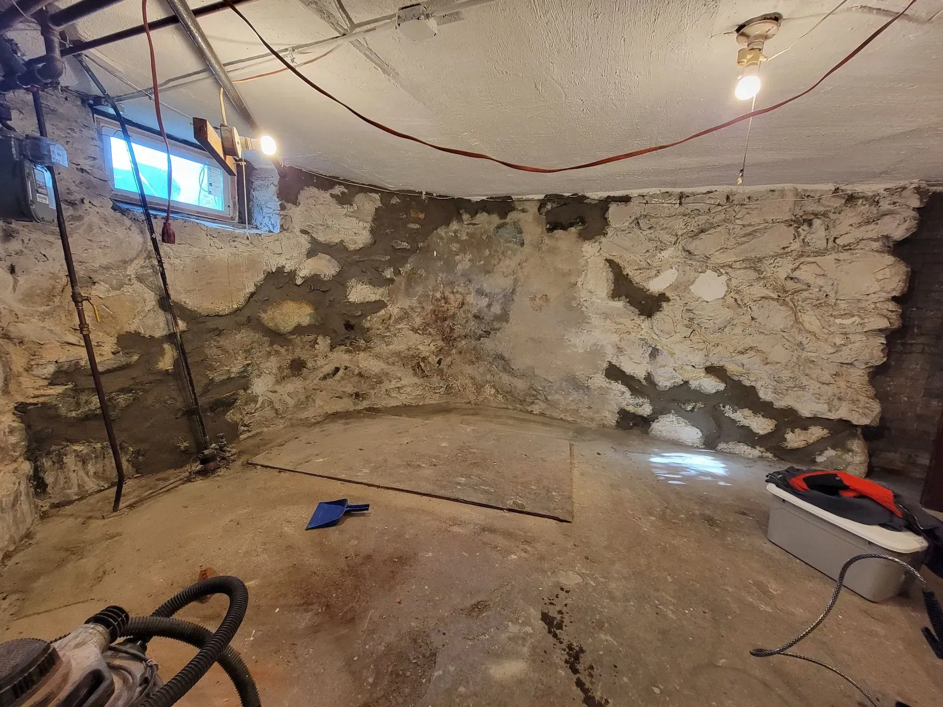 Basement corner with exposed stone walls, partially plastered. Concrete floor, overhead lights, and tools.
