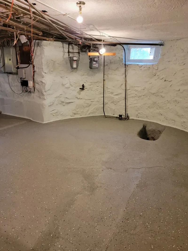 Basement with concrete floor, white stone walls, exposed pipes and electrical, small window.
