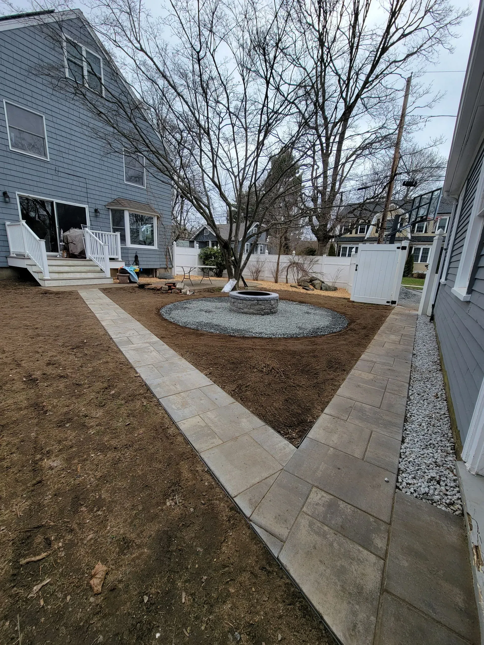 Backyard with concrete walkways leading to a fire pit surrounded by mulch and rocks, near a house.