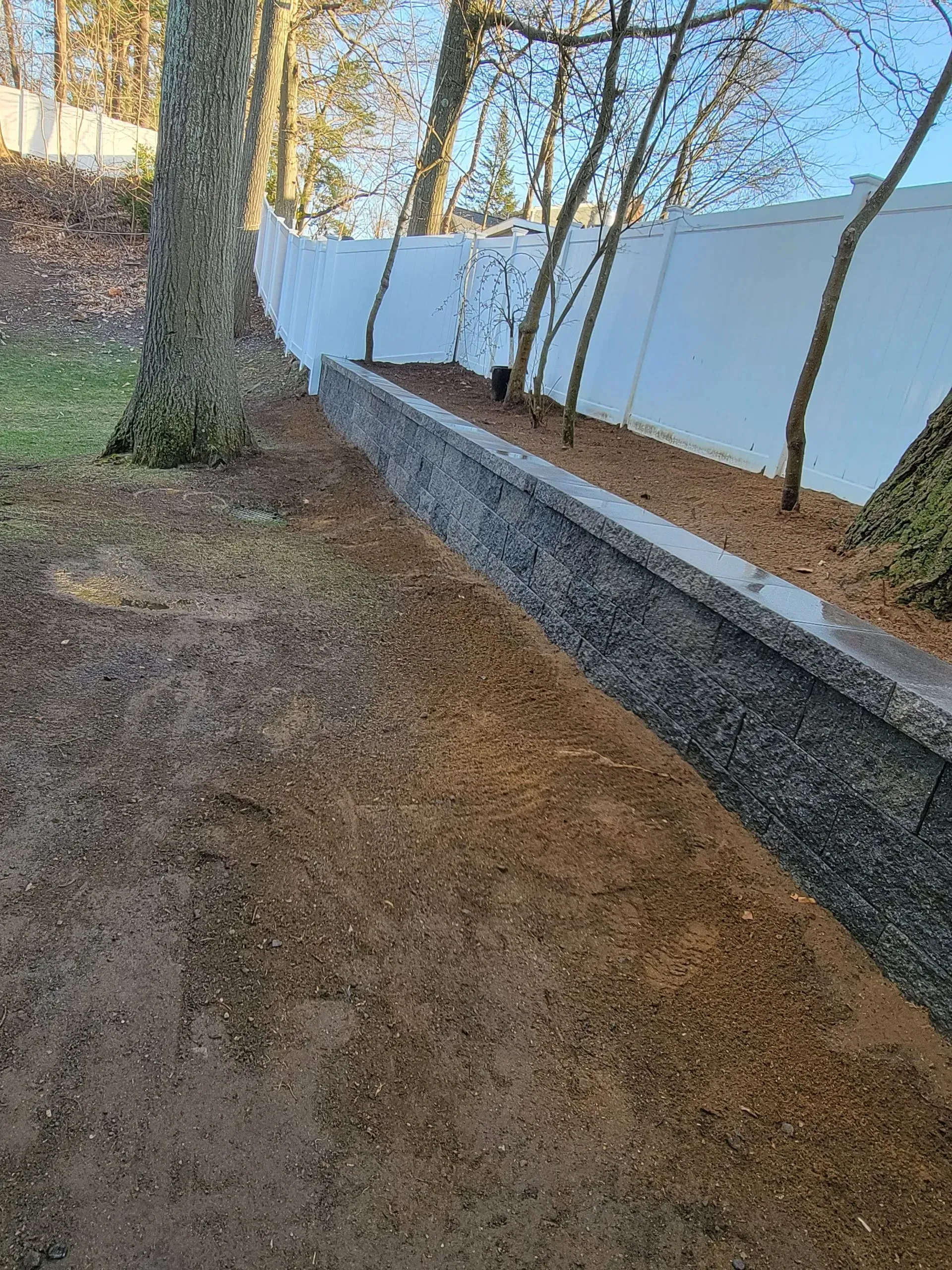 A retaining wall of dark blocks with a light-colored top, bordered by mulch and a white fence.