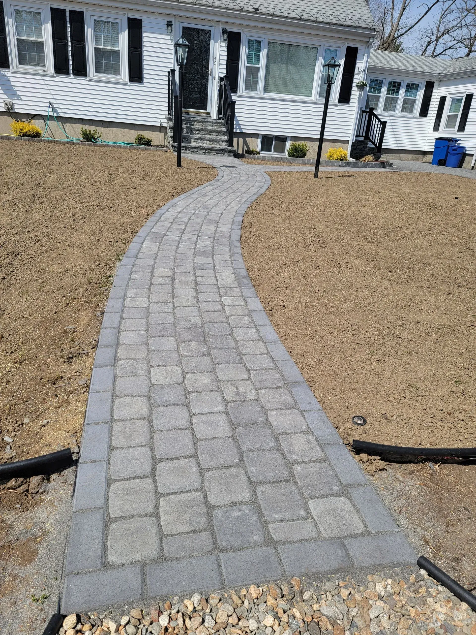 Brick pathway leading to a white house with black shutters, on a brown gravel lawn.