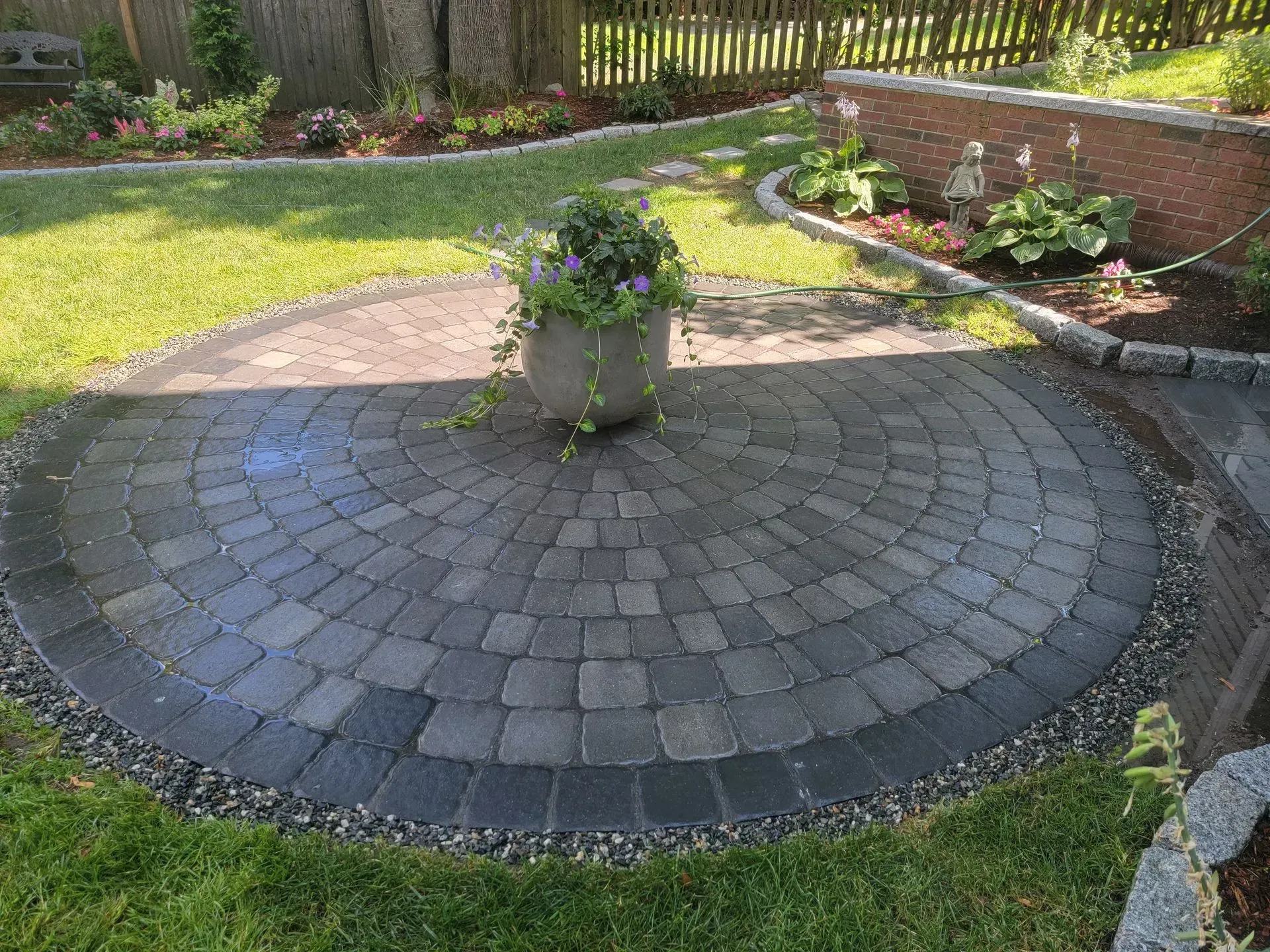 Circular paved patio with potted flowers, surrounded by grass and flower beds.