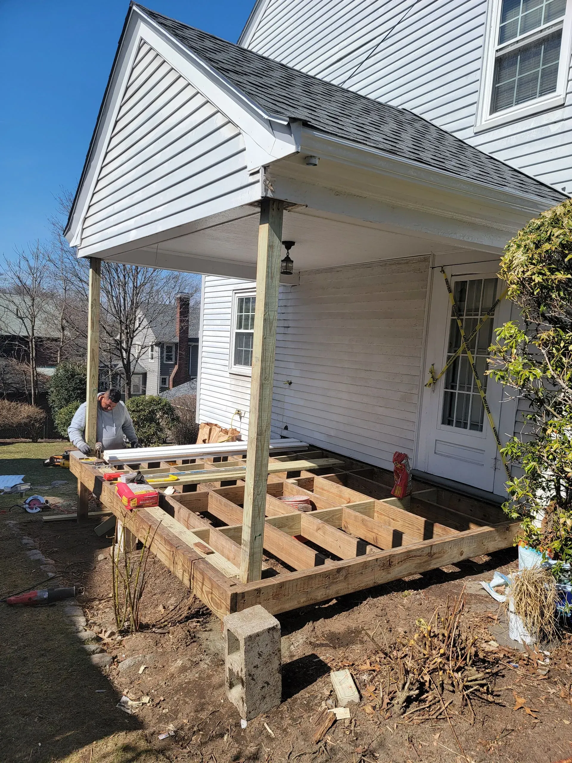 A partially built wooden deck with a man working on it, attached to a white house with a covered porch.