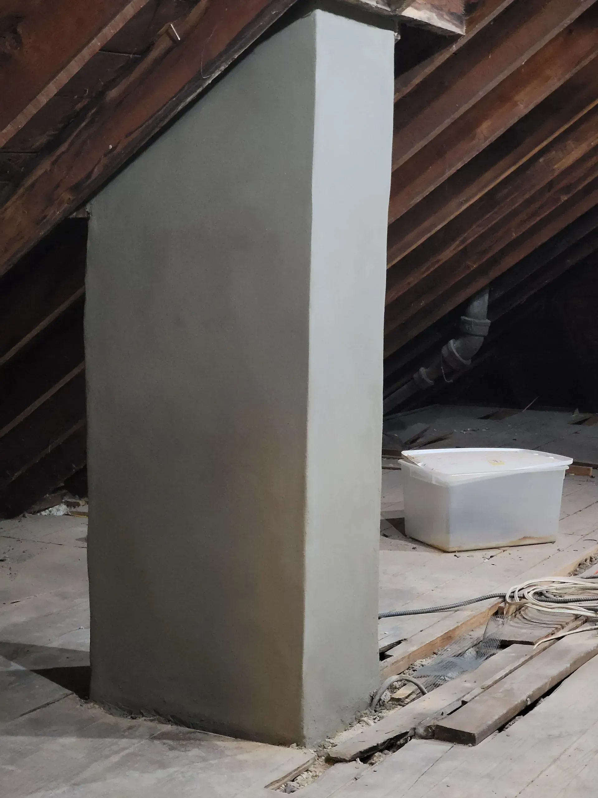 A gray, rectangular chimney in an attic with wooden rafters. A white bucket sits nearby.