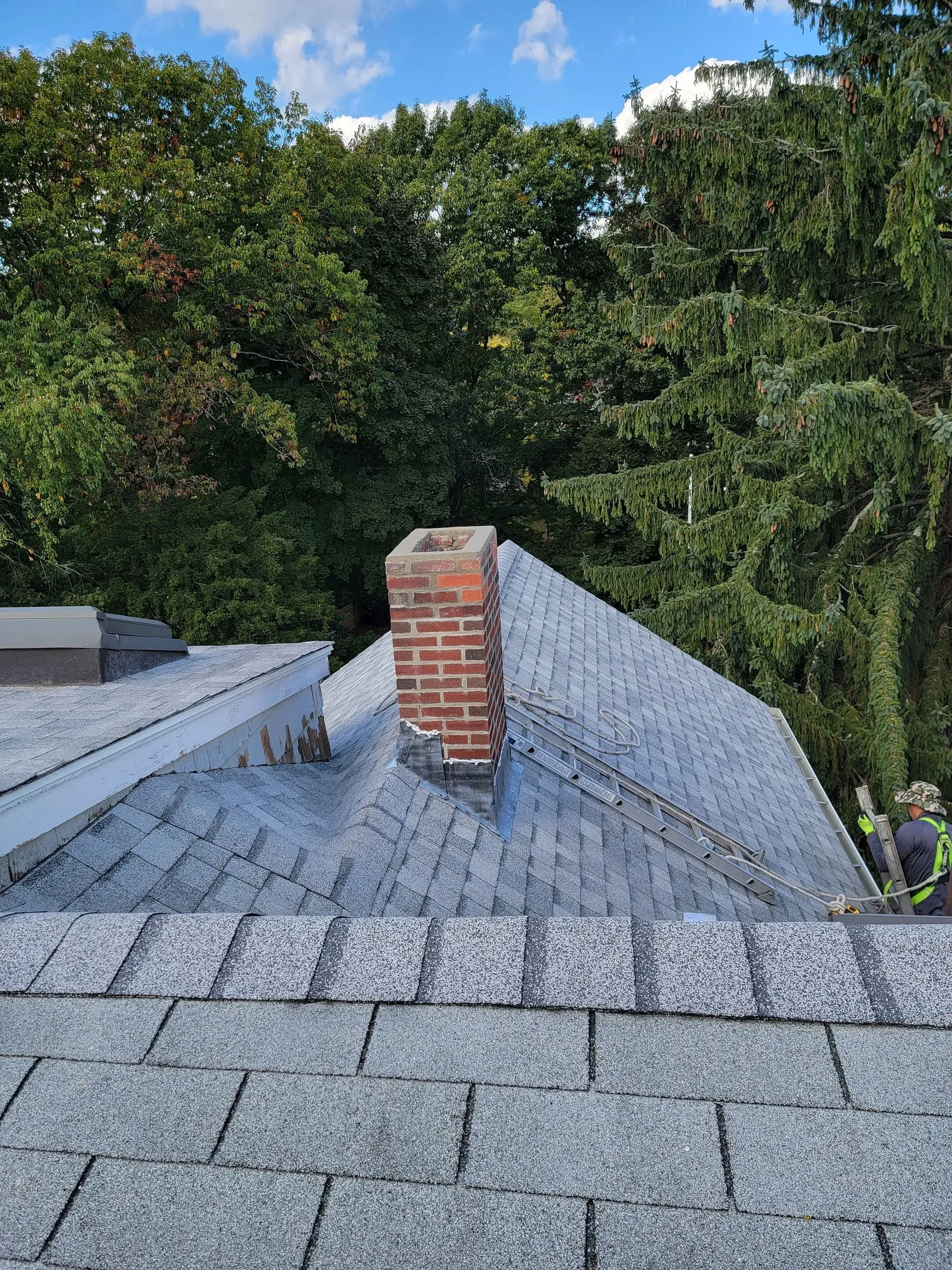 Rooftop with a brick chimney and new gray shingles, with a person wearing safety gear on the side.