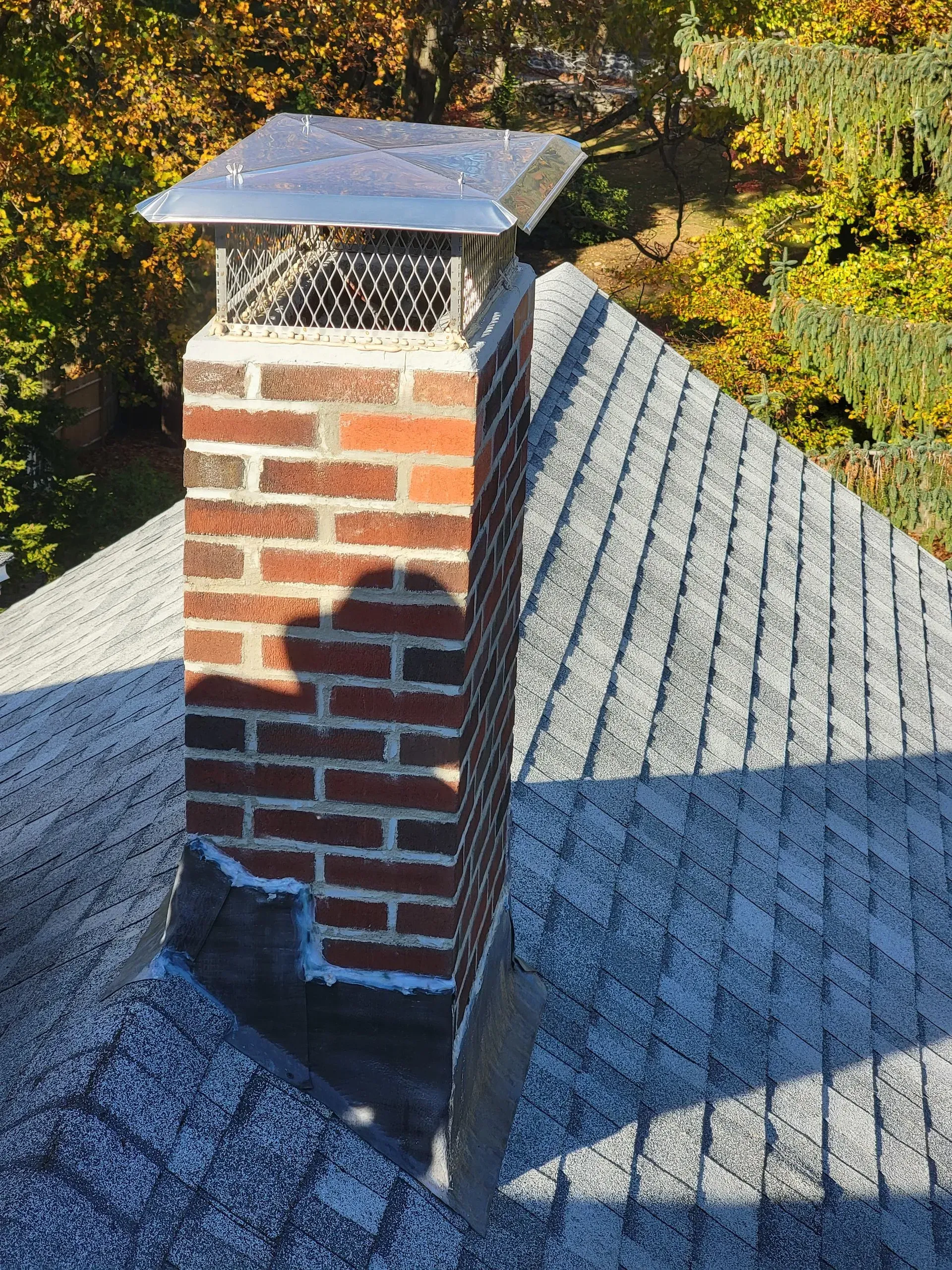 Brick chimney with cap on a gray shingled roof, casting a shadow.