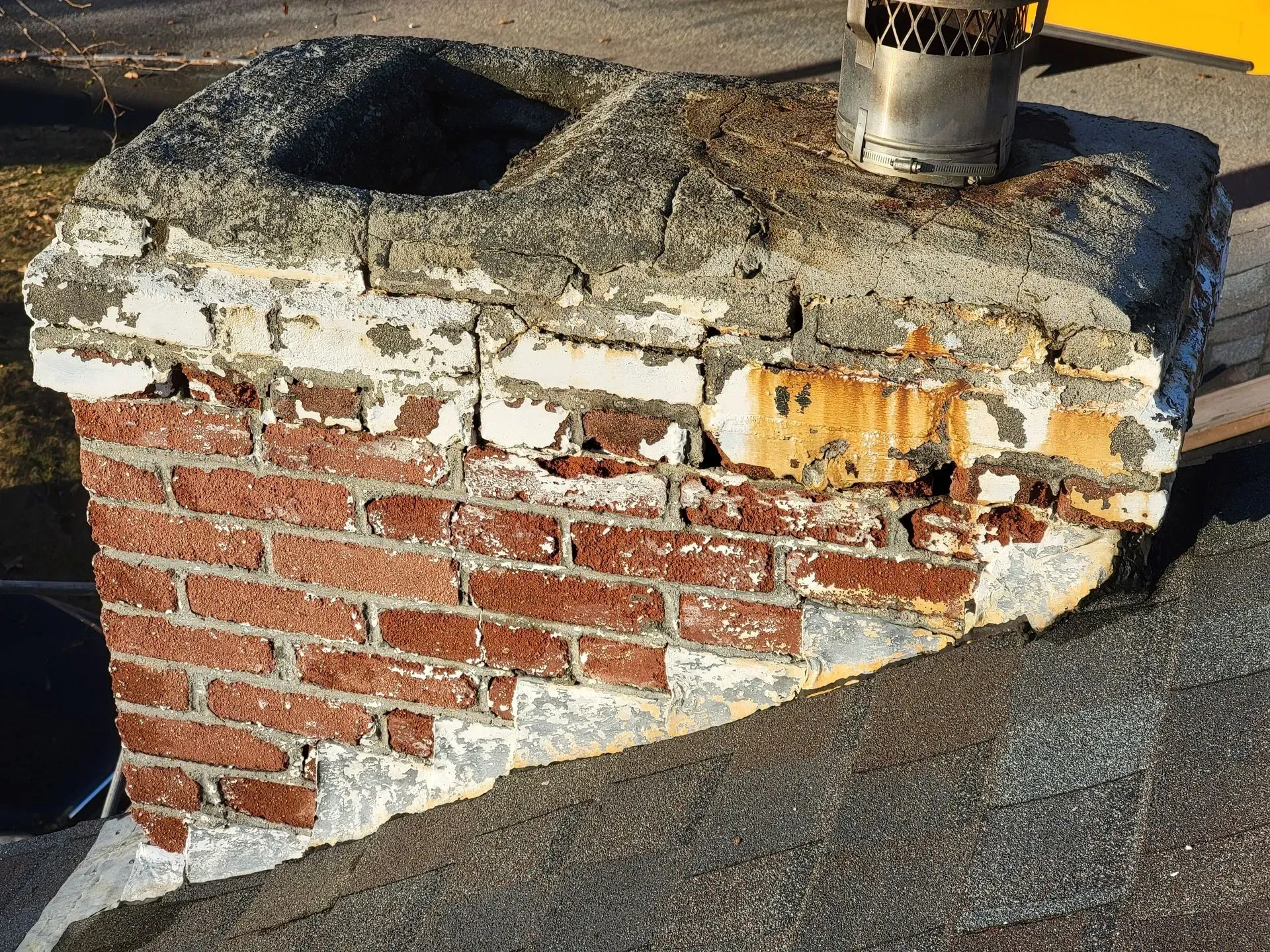 Damaged brick chimney with deteriorated concrete cap and flue, on a rooftop.