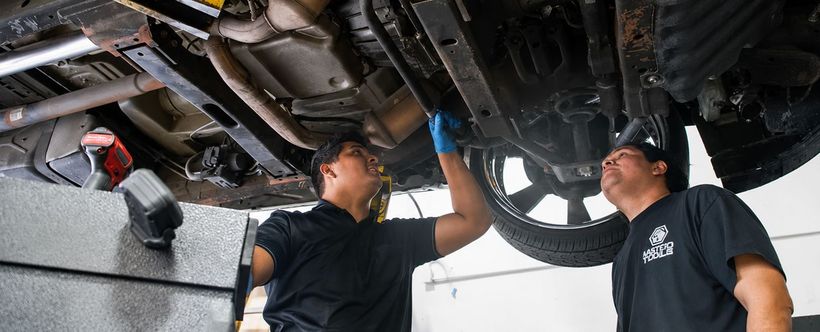 Two mechanics inspecting the underside of a vehicle in a garage, one pointing at a part overhead.
