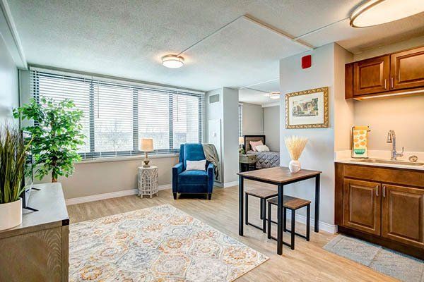 Bright, furnished studio apartment with seating area, kitchenette, and bedroom visible in the background.
