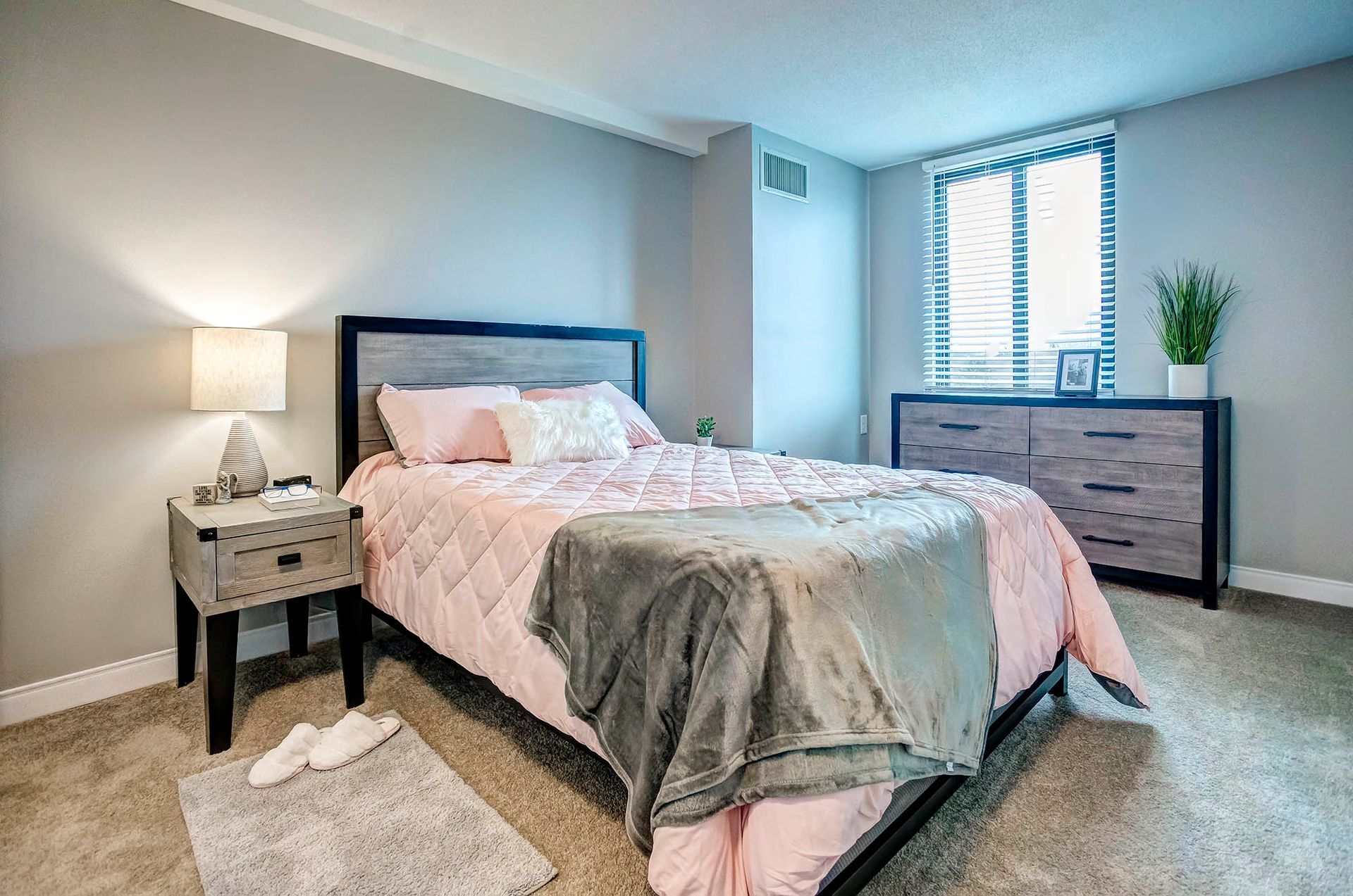 Bedroom with a bed, nightstand, dresser, and window. Gray walls, pink bedding, and beige carpet.