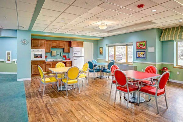 Dining area with colorful chairs, wooden floor, kitchen, and large windows.