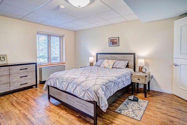 Bedroom with wooden floor, bed, dresser, nightstand, window, and rug.