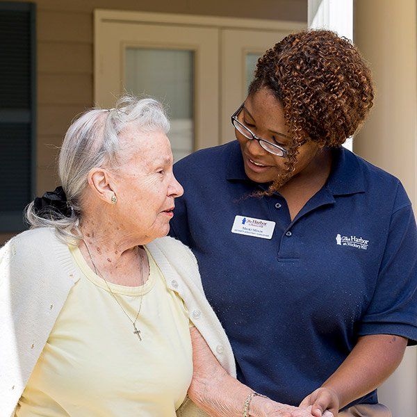 Woman in blue shirt assisting an elderly woman wearing a cardigan. Outdoors, porch setting.