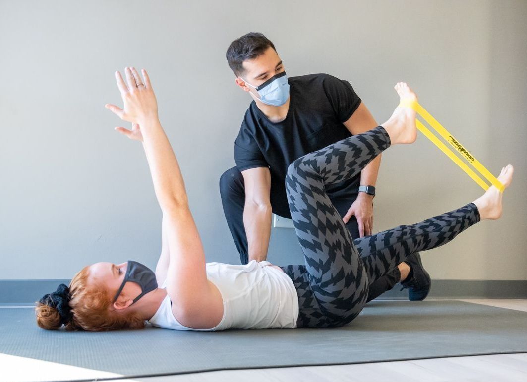 A physical therapist guides a person lying on a mat as they perform a leg exercise using a yellow resistance band.