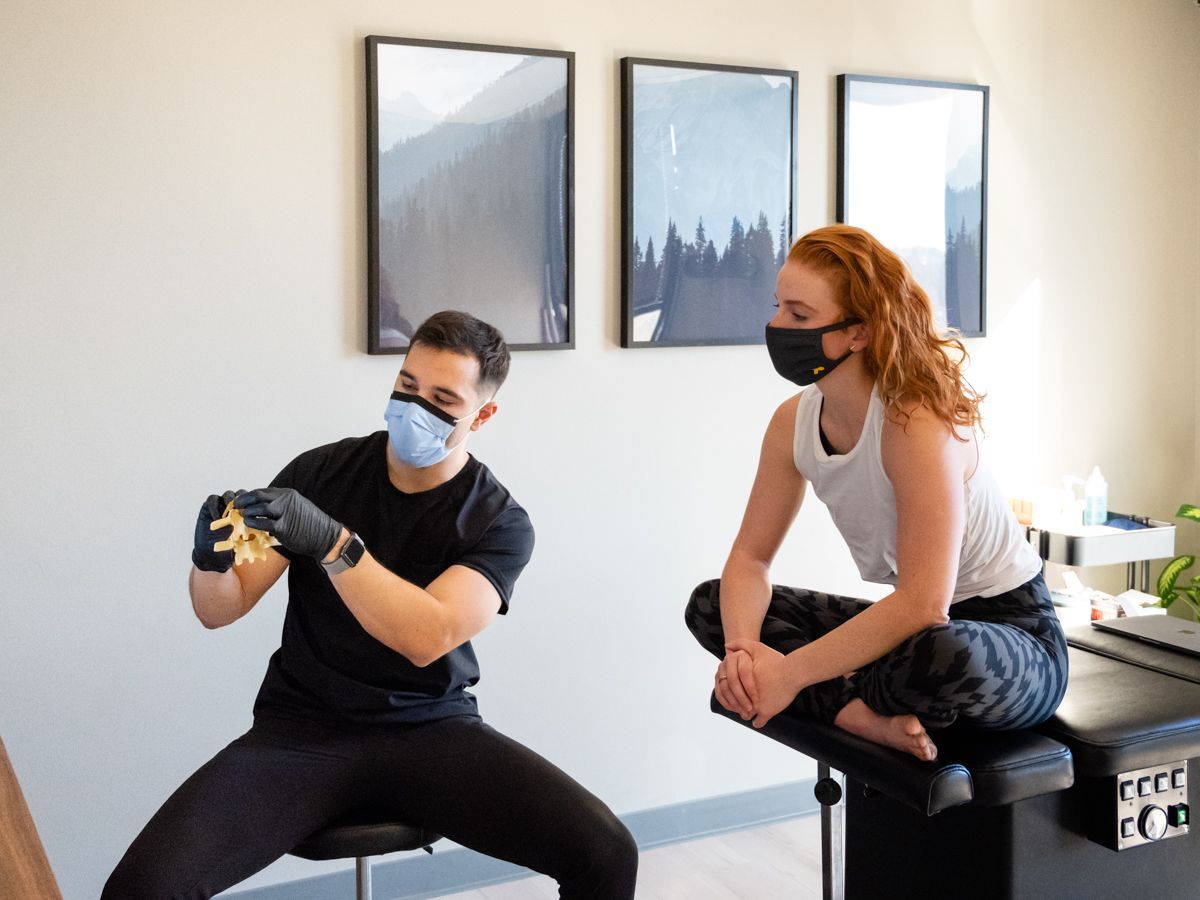 A practitioner in a black shirt and face mask holds a spinal model to explain it to a patient sitting on an exam table.