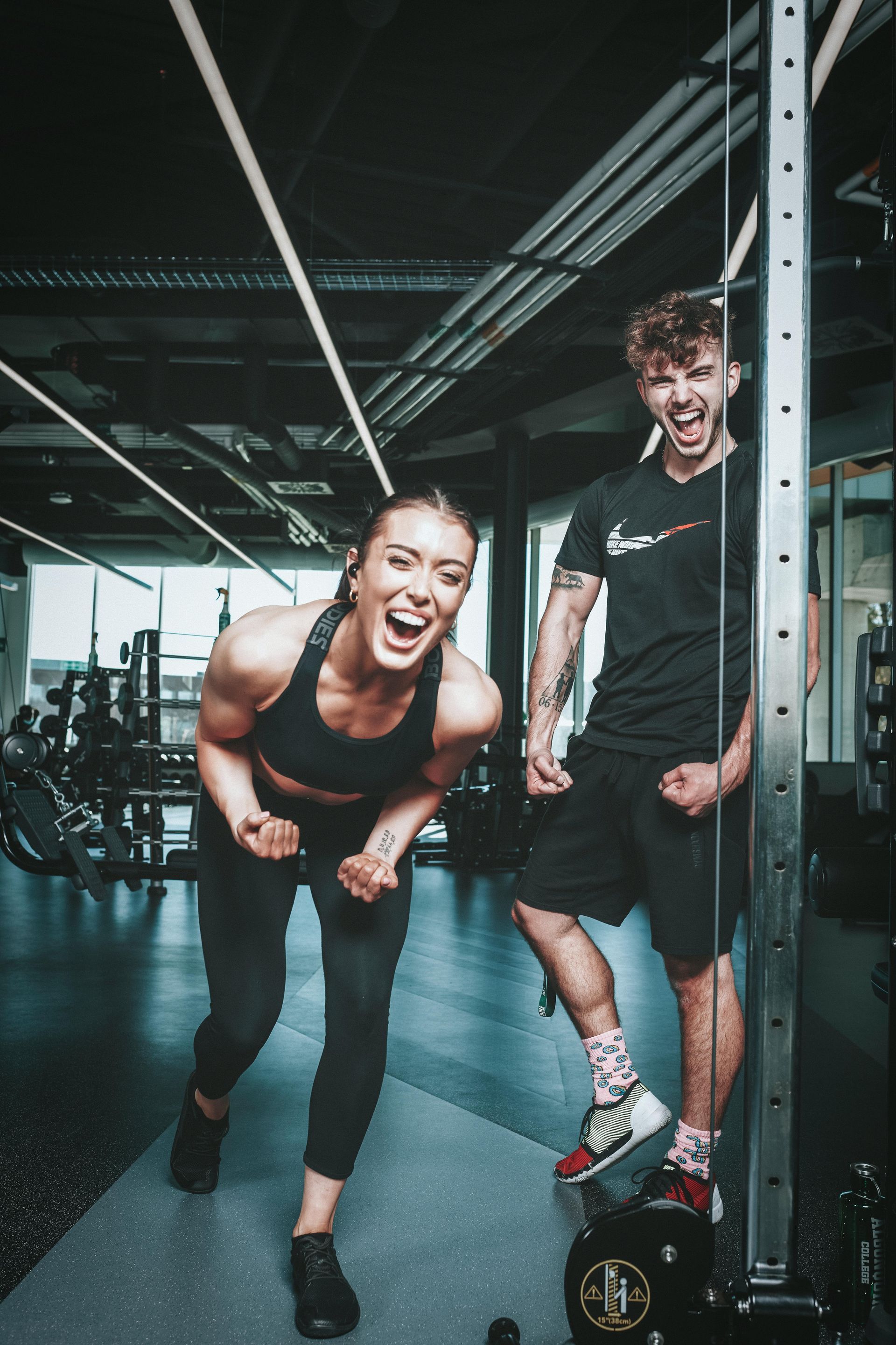 Two people in a gym laughing and posing excitedly, wearing athletic clothes, with weightlifting equipment in the background.