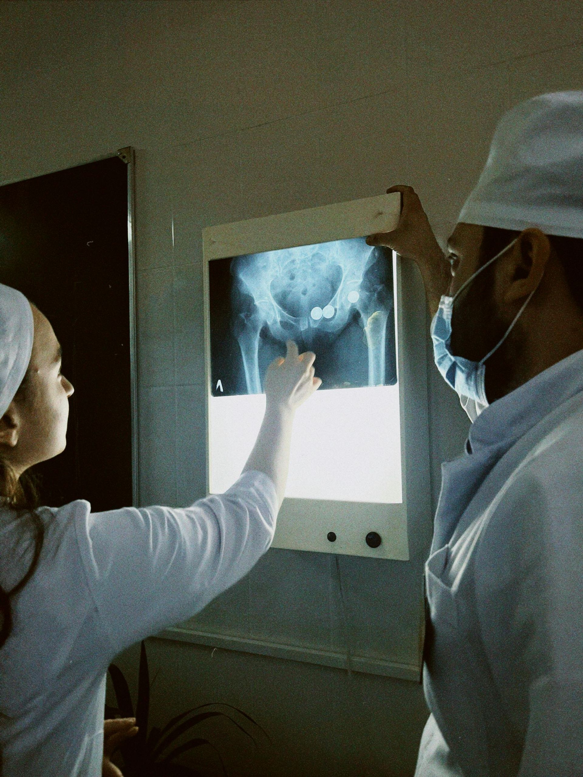 Two medical professionals in white coats and caps examine an X-ray of a pelvis on a lightbox.