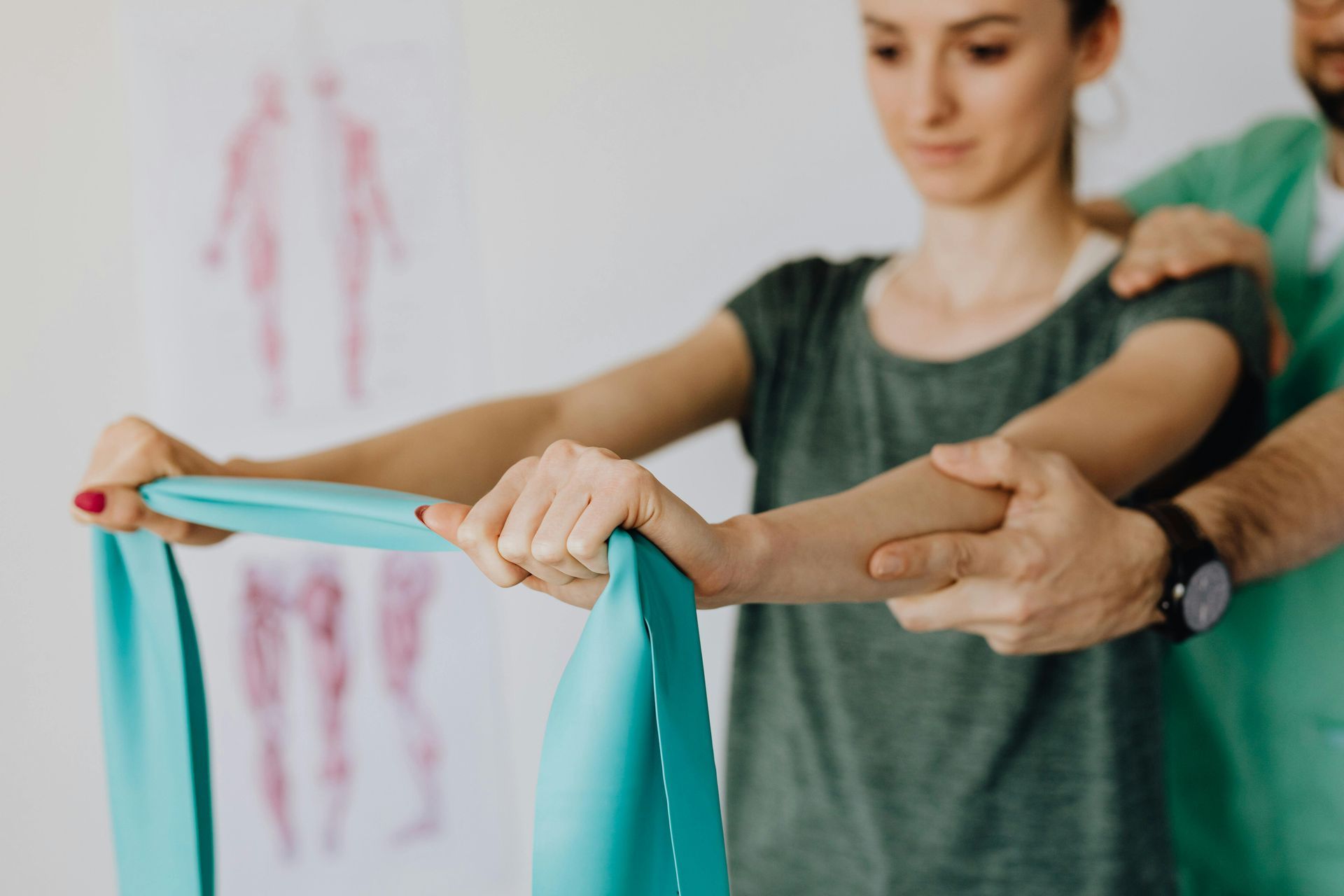 A physical therapist guides a patient in performing an arm exercise using a teal resistance band in a clinical setting.