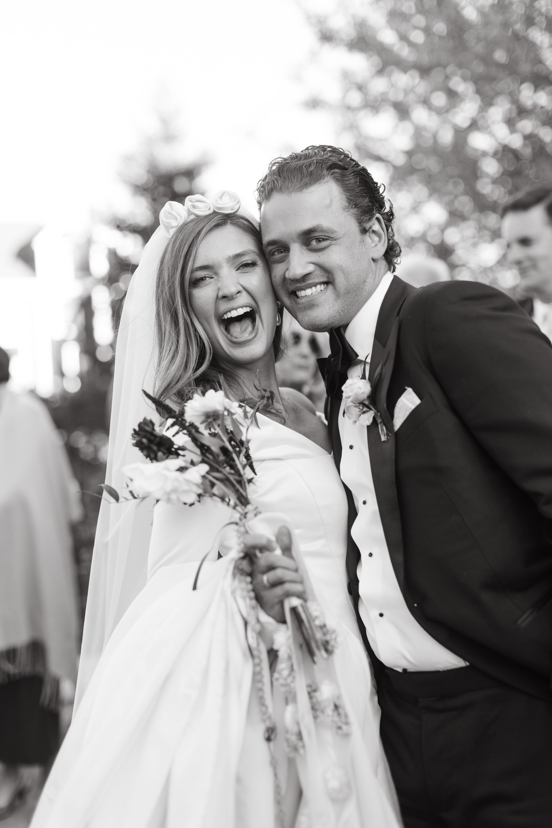 Bride and groom laugh at wedding; black and white photo. Bride wears gown, veil, and holds bouquet. Groom in tuxedo.