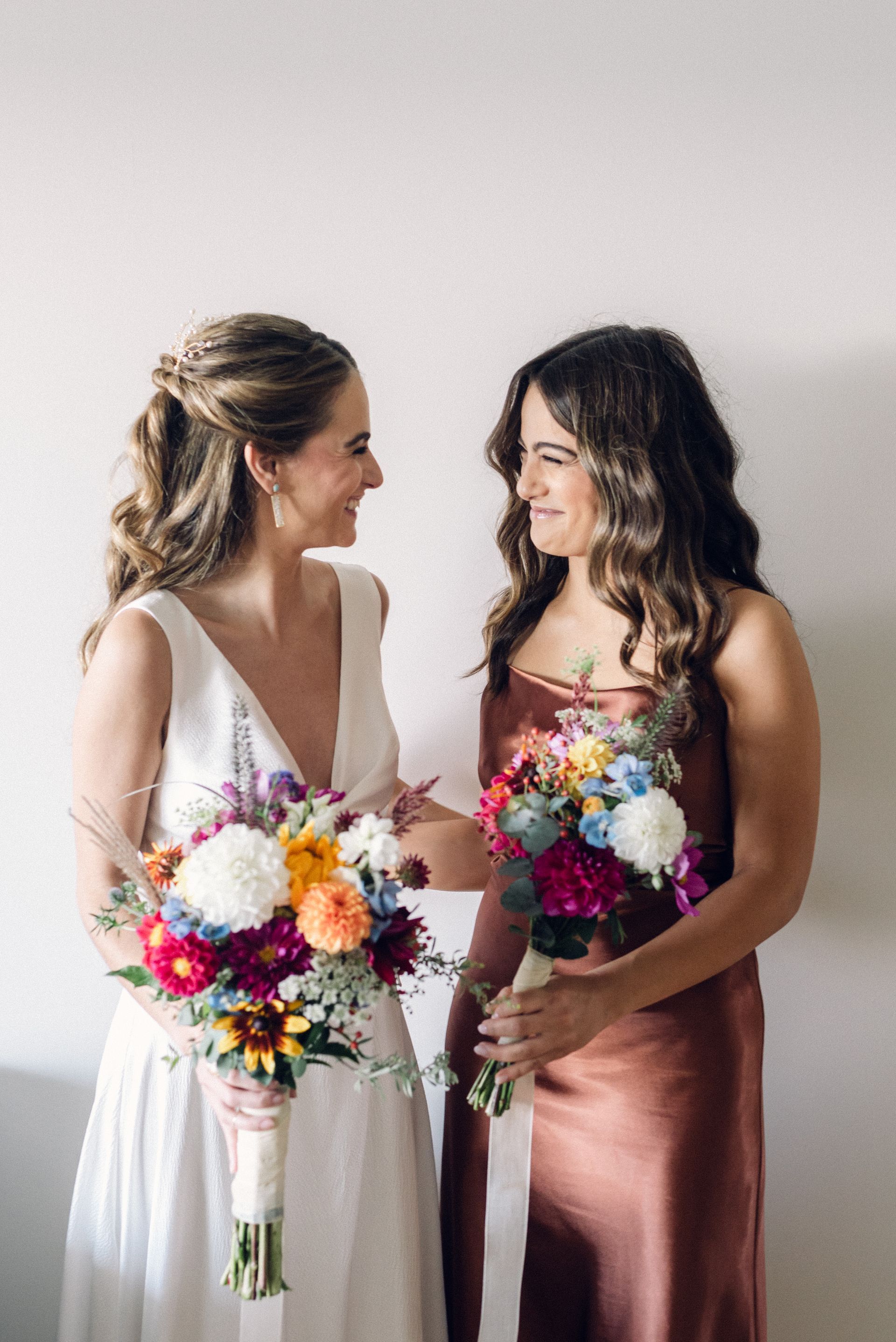 Bride and bridesmaid smile, holding bouquets. White dress, copper dress, soft light, white background.