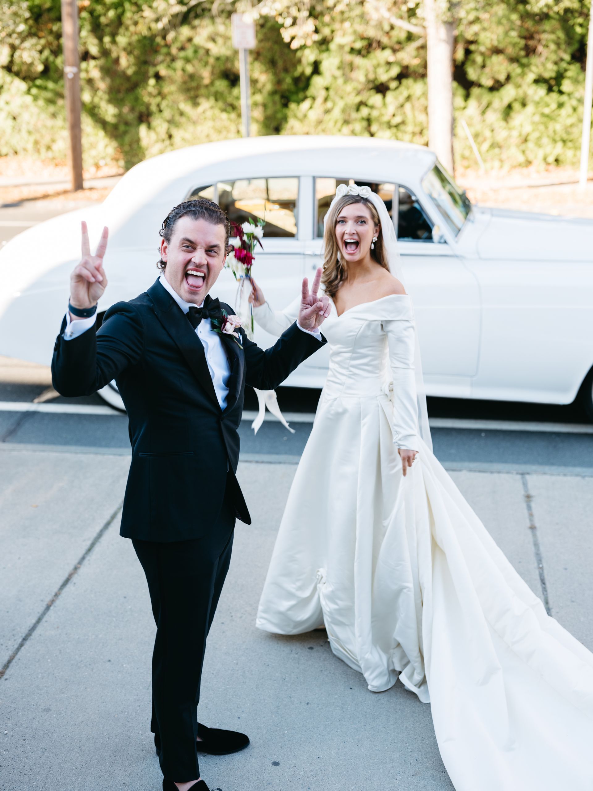 Newlyweds smile and give peace signs; classic white car in background.