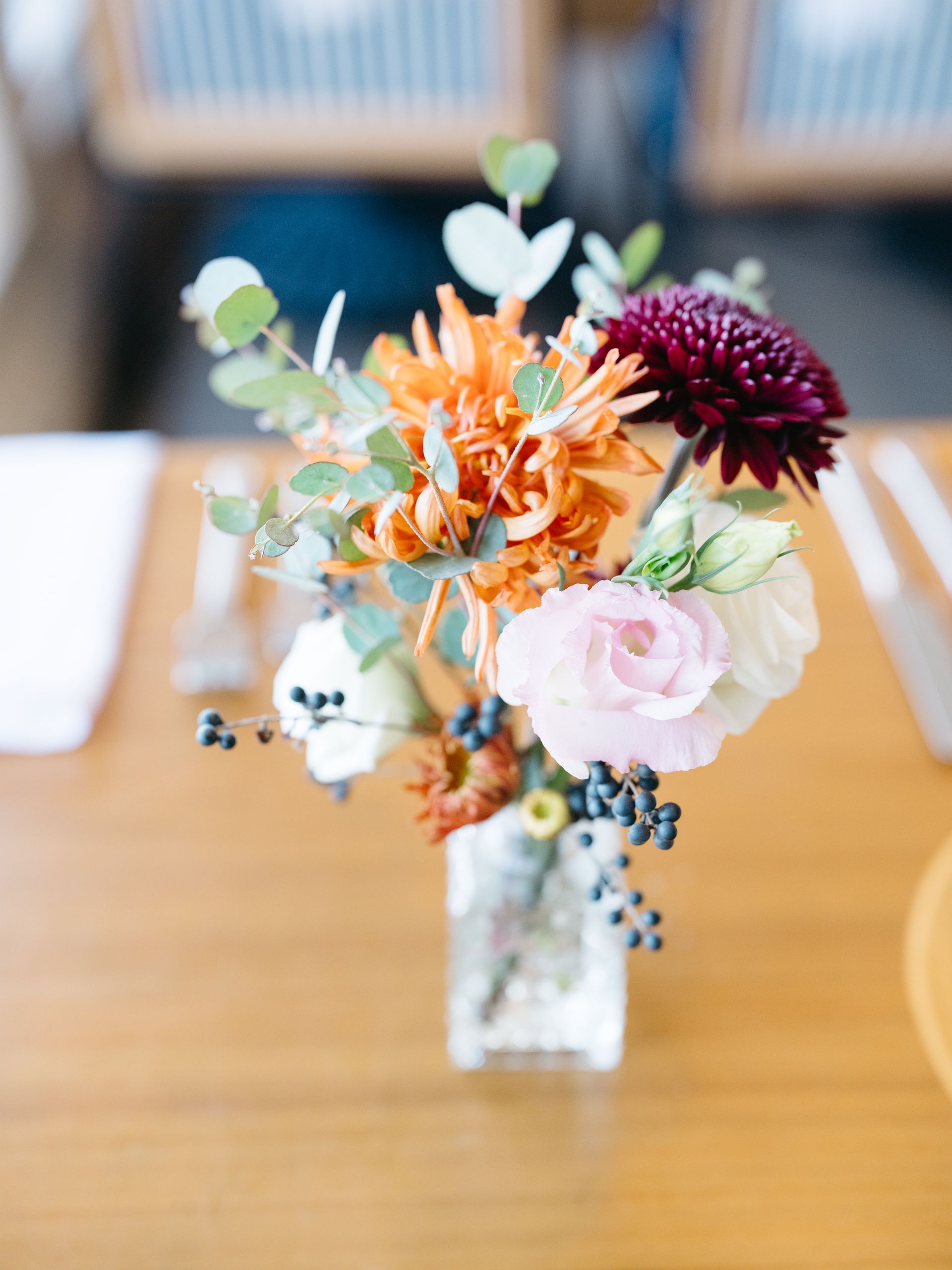 Close-up of a small floral arrangement in a clear square vase on a wooden table with silverware.
