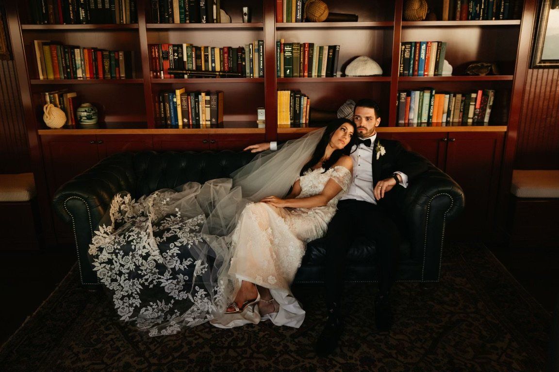 Newlyweds on green sofa in library. Bride in lace gown, groom in tuxedo.