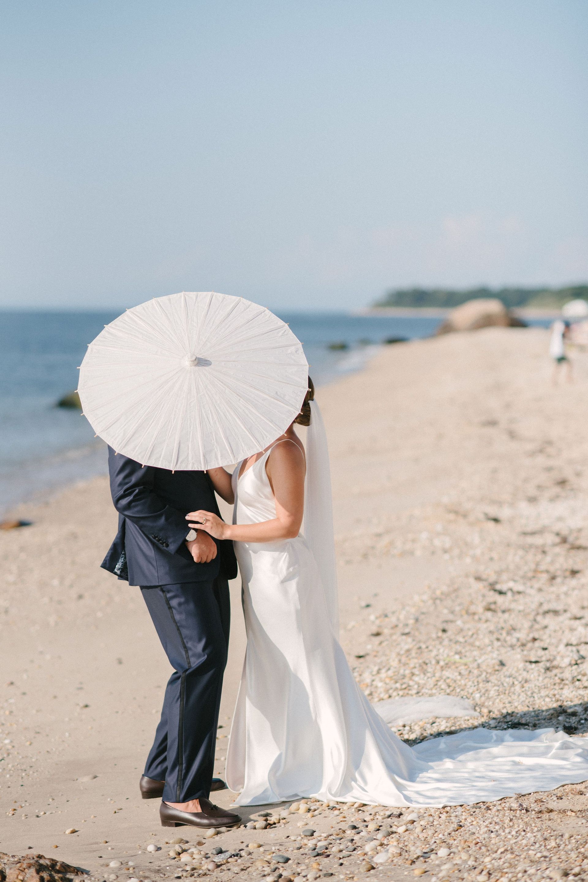 Bride and groom on a beach, hidden under a white parasol, kissing.