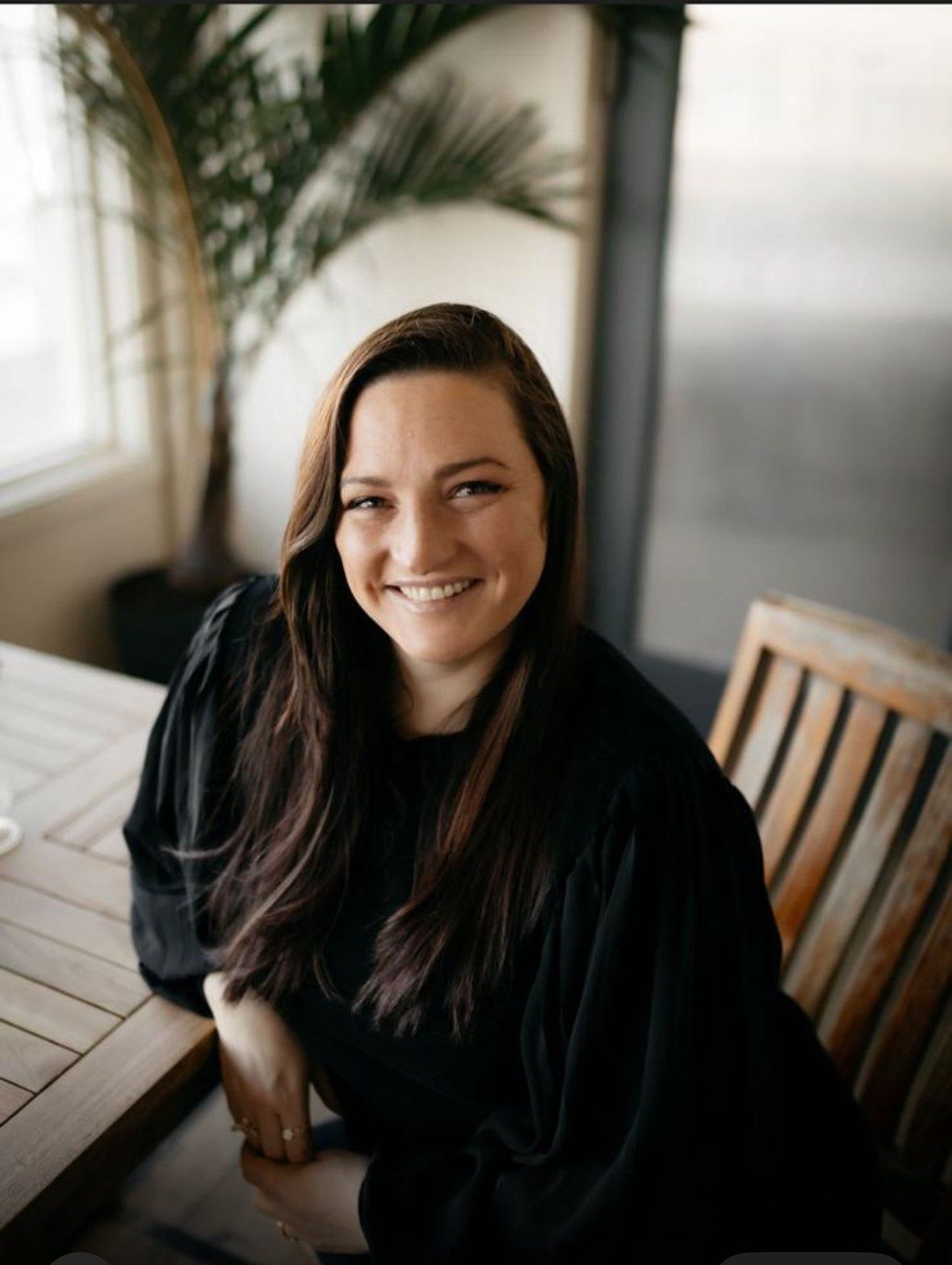 Woman with long brown hair smiles while sitting at a wooden table, indoors.