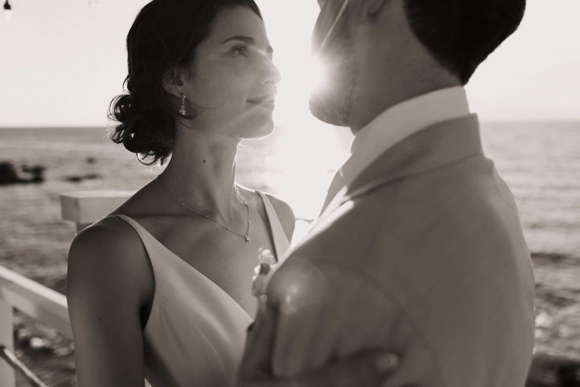 Couple embraces on a pier at sunset, the woman looks up, sun flares between them.