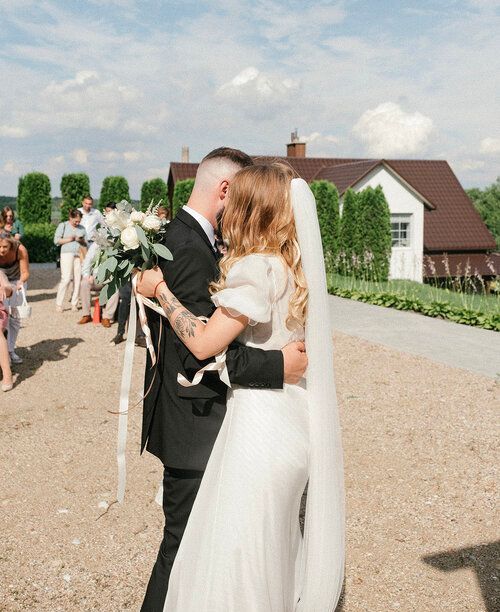 Newlyweds embrace outdoors after their wedding ceremony. Bride in white gown, groom in black suit, guests in background.