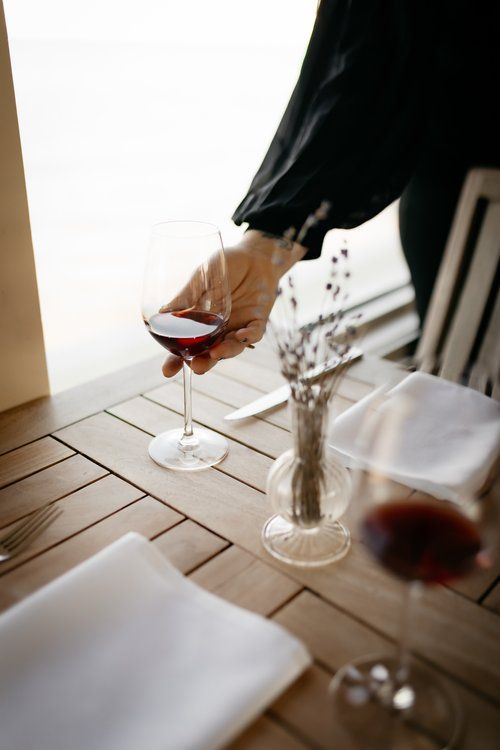 Person holding a glass of red wine at a restaurant table set with a napkin, fork, and vase of flowers.