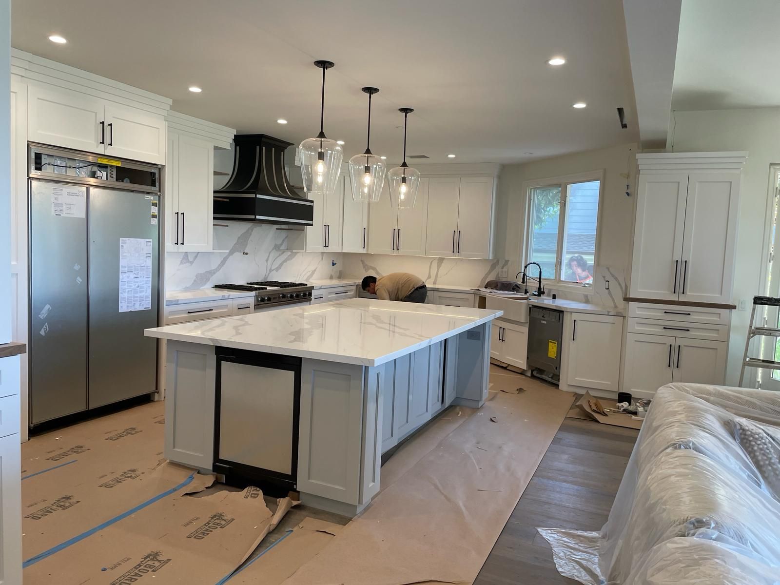 Modern kitchen under construction with white cabinets, large island, and stainless steel appliances.