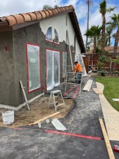 House exterior under construction, with a worker applying stucco. Windows taped, tools and materials present.