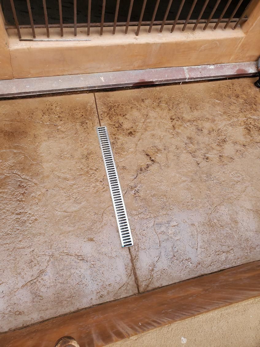 Close-up of a concrete surface with a metal grate drain, near a wall and a window with bars.