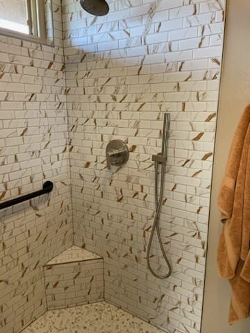 Shower with white and gold-toned brick-like tiles, a handheld showerhead, and a grab bar.