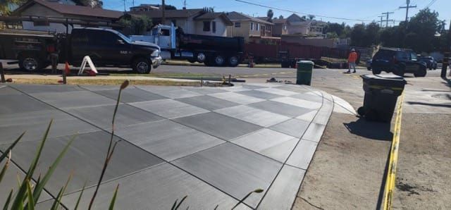 Concrete checkered sidewalk next to a curved curb. Trucks and buildings in the background on a sunny day.