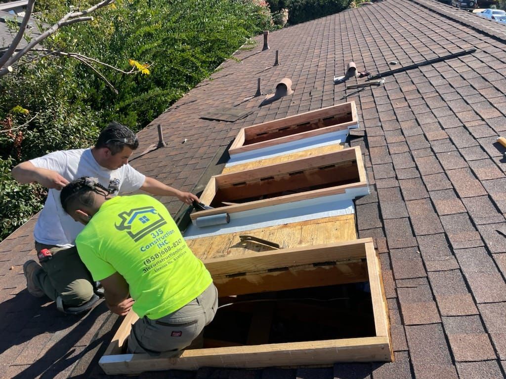 Two workers installing skylights on a shingled roof under sunny skies.