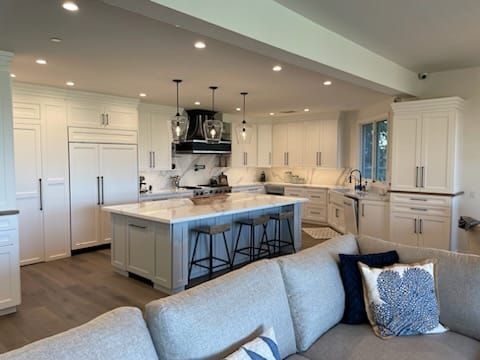 Spacious, white kitchen with island, pendant lights, and stainless steel appliances; a gray couch is in the foreground.
