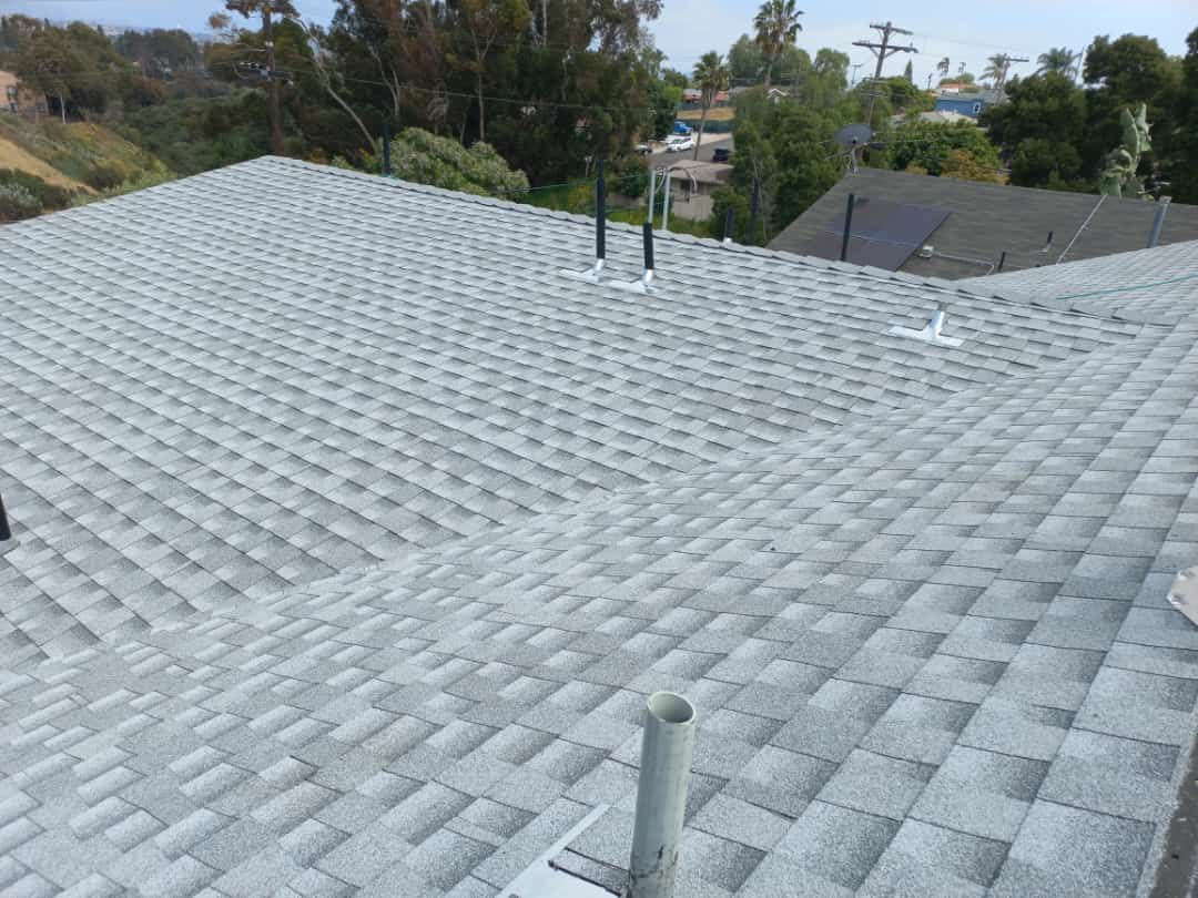 Gray asphalt shingle roof, angled view. Several vents and pipes. Sunny outdoor setting.
