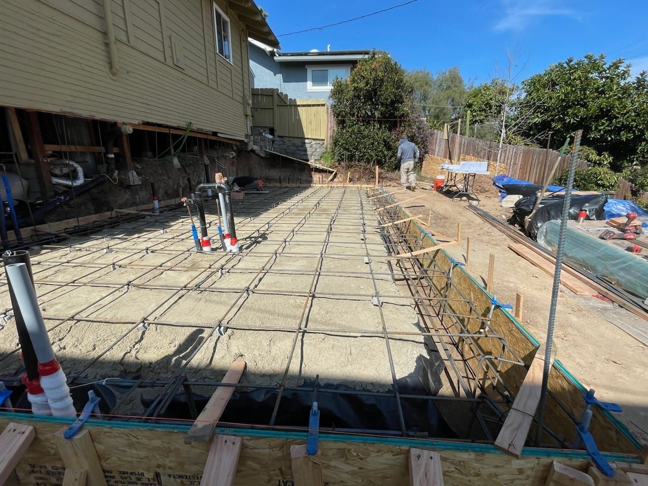 Construction site: Rebar grid laid over sand, wooden forms, and workers in the background.