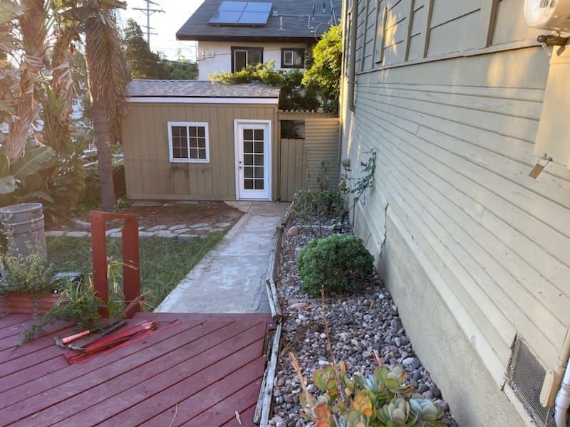 A narrow concrete path leads to a tan shed with a white door, beside a light-colored house.