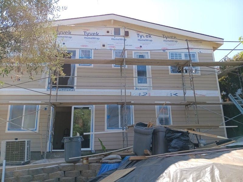 Two-story house under construction with beige siding, scaffolding, and windows.